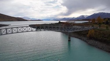 Havadan çekilen görüntülerde Tekapo Gölü 'nün sakin manzarası, bir köprü, turkuaz sular ve dağları çevreleyen yumuşak, dağınık ışık altında yer alır.