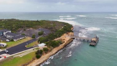 Drone, Port Campbell 'ın engebeli kıyı şeridini, rıhtımını ve bulutlu gökyüzünün altındaki yemyeşil çimlerini görüntülüyor.