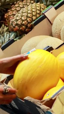 Kanarya kavunlarını canlı bir süpermarket ortamında inceleyen bir insan, doğal ışıklandırmayla taze ürünleri ön plana çıkarır.