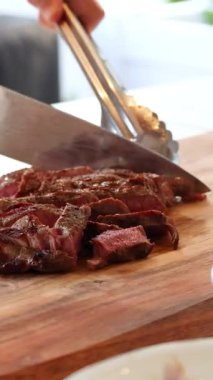 Bir şef ahşap bir tahtada pişmiş bifteği ustalıkla dilimliyor, iyi aydınlatılmış bir mutfakta mutfak becerileri sergiliyor.