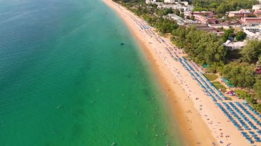 Hava görüntüleri canlı bir Phuket sahili yakalıyor turkuvaz suları, güneş banyosu ve şemsiyelerle, canlı bir tatil yeri atmosferi sergiliyor.