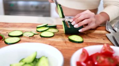 Parlak bir mutfakta ahşap bir tahtanın üzerinde salatalık dilimleyen eller. Taze malzemeler ve doğal ışıklandırma temiz, davetkar bir atmosfer yaratır.