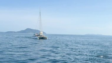 Bir katamaran Chalong Körfezi, Phuket 'te, açık gökyüzünün altında görünür adalarla sakince yelken açar. Sahne, dingin okyanus güzelliğini yakalar