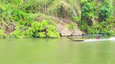 Uzun kuyruklu bir tekne Kanchanaburi, Tayland 'daki Kwai nehri boyunca hızla ilerliyor, yemyeşil ve sakin sularla çevrili.