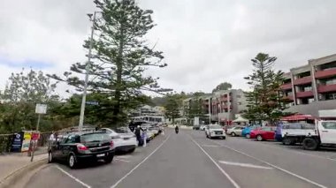 Büyük Okyanus Yolu 'ndaki kalabalık bir caddede bir gezinti, park halindeki arabaları, yayaları ve bulutlu gökyüzünün altındaki yemyeşil alanları gösteriyor.