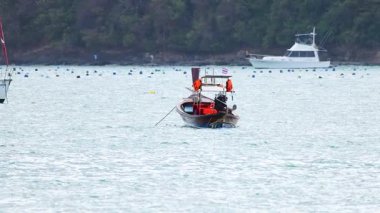 Uzun kuyruklu bir tekne Tayland, Phuket yakınlarındaki sakin sularda yol alır. Sahne, sakin okyanus manzaralarını ve yumuşak ışık altında uzak yatları yakalıyor.