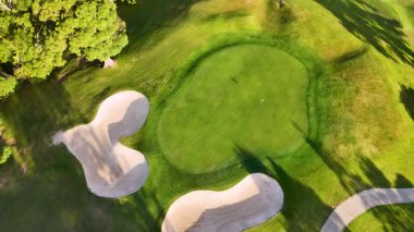 İnsansız hava aracı, altın saat aydınlatması altında sığınakları ve yeşili olan bir golf sahası yakalıyor, gölgeleri ve manzarayı renklendiriyor.