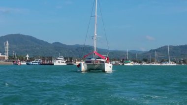 Tayland, Phuket yakınlarındaki turkuaz sularda bir katamaran yelken açıyor. Dağlık bir zemin ile berrak gökyüzünün altında.