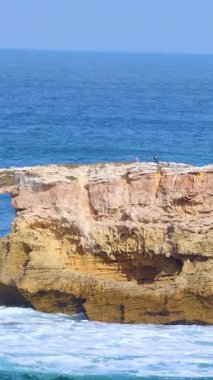 Okyanus dalgaları, berrak gökyüzünün altındaki engebeli kayalıklara çarpar ve su ile kayanın dinamik etkileşimini yakalar.