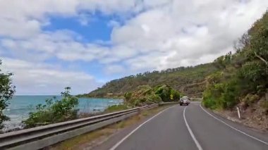 Parlak bulutlu bir gökyüzü altında, yemyeşil ve kıyı manzaralarıyla çevrili, muhteşem Büyük Okyanus Yolu boyunca bir araç seyahat eder.