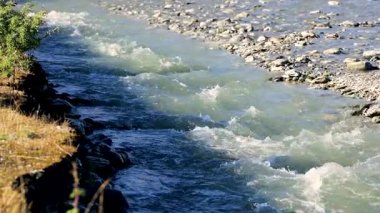 Parlak güneş ışığı altında kayalık bir arazide akan sakin bir nehir doğal güzellik ve sükuneti gözler önüne seriyor.