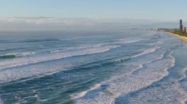 Havadan çekilen görüntüler dalgaların yumuşak sabah ışığı altında sakin bir sahile yuvarlandığını gösteriyor. Uzak binalar görülebilir.