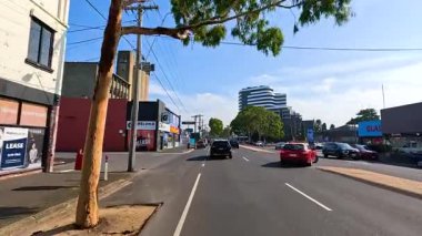 Geelong 'da gündüz araba kullanmak, şehir binalarını, trafiği ve açık gökyüzünü göstermek. Dinamik sokak manzaralarını ve canlı şehir yaşamını yakalar