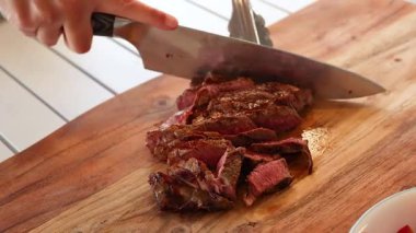 Bir insan iyi aydınlatılmış bir mutfak ortamında ahşap bir tahtada pişmiş biftek dilimliyor.