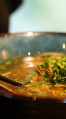 Bangkok restoranındaki bir kase çorbadan erişte aşıran yemek çubuklarının yakın çekimi. Ilık aydınlatma canlı renkleri güçlendirir
