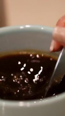 Bir el mavi bardakta kahve karıştırıyor, dalgalar ve kabarcıklar yaratıyor. Ilık aydınlatma sıcak atmosferi güçlendirir