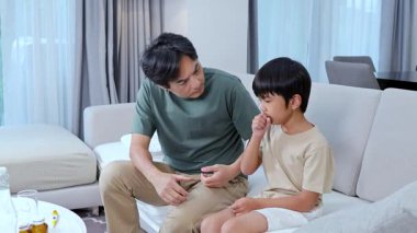 Bir baba hasta çocuğuna kanepede bakar, sıcaklığı kontrol eder ve parlak bir oturma odasında rahatlık sunar.