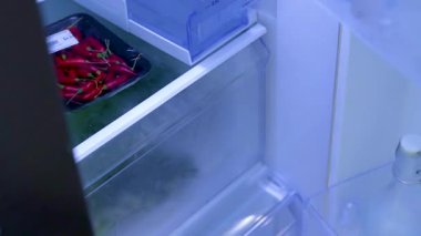 Bir insan buzdolabını açar ve mutfak ortamında parlak ışık altında taze sebzeler seçer.