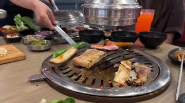 Canlı bir Bangkok restoranında barbekü masasında ızgara domuz eti ve sebze. Renkli yemekler ve içeceklerle dolu canlı bir atmosfer.