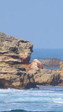 Dalgalar, açık mavi gökyüzü altında engebeli uçurumlara çarpar ve deniz ile kaya arasındaki dinamik etkileşimi gösterir.