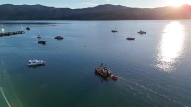 Gün batımında Akaroa Limanı 'ndaki teknelerin hava görüntüsü, sıcak güneş ışığı altında sakin suları ve manzaralı tepeleri yakalıyor.