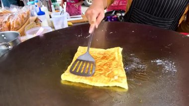 Bir satıcı, Tayland pazarında parlak bir ışık altında sıcak bir ızgarada ustalıkla altın-kahverengi bir roti çevirir.