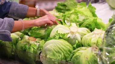 Eller, iyi aydınlatılmış bir süpermarket ortamında taze ürün seçimini ön plana çıkaran marul başlarını dikkatle topluyor.