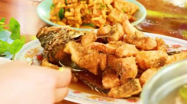 Sıcak bir ortamda, canlı Tayland yemekleri ile çevrili soslu kızarmış balık servis eden bir el.