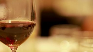 Yumuşak ışıklandırılmış yemek ortamında kırmızı şarap kadehinin yakın plan çekimi, bulanık bir arka plan, sıcak ve samimi bir atmosfer yaratır.