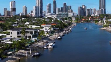 İnsansız hava aracı görüntüleri tekneli sakin bir kanal yakalıyor ve açık mavi gökyüzünün altında Altın Sahil silueti yükseliyor.