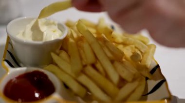 Bir el kızartmayı mayoneze batırır ve yumuşak ışığın altında çizgili bir tabağa ketçap sürer.