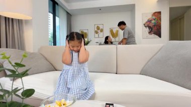 Modern bir oturma odasında ebeveynler tartışırken genç bir kız koltukta kulaklarını kapatıyor.