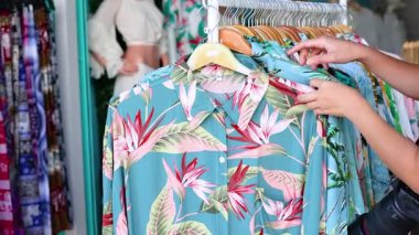 Bir insan, iyi aydınlatılmış bir pazar ortamında, askıdaki canlı çiçekli gömlekleri inceler.