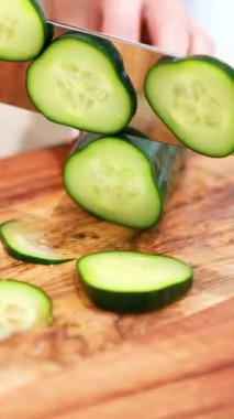 Ahşap bir tahtanın üzerinde salatalık kesen ellerin yakın çekimi. Parlak ışıklandırma, taze ürün ve keskin bıçak becerilerini ön plana çıkarıyor