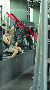 Spor salonunda oturan bir adam, akıllı telefonuna odaklanmış, egzersiz ekipmanları ve diğer spor salonlarına gidenlerle çevrili.