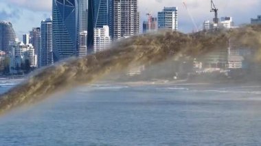 Kum, açık gökyüzü altında kıyı erozyonu yönetimini gösteren bir şehrin ufuk çizgisi yakınındaki okyanusa pompalanır.