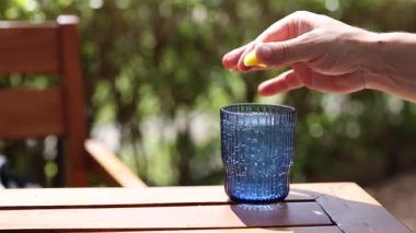 Bir el, güneşli ahşap bir masada mavi bir bardağa limon suyu sıkıyor ve canlandırıcı bir sahne yaratıyor.