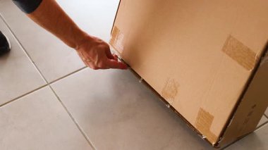 Bir insan fayanslı zeminde bantlanmış bir karton kutuyu açmak için bıçak kullanır.
