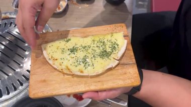 Bir el, Bangkok 'ta bir restoranda erimiş peyniri bir tabağın üzerine koyuyor, Kore barbekü seti ve canlı ışıklandırmayla vurguluyor.