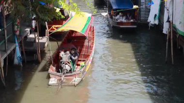 Bir kano, Bangkok 'taki Khlong Lat Mayom Yüzen Pazarı' nda, parlak gün ışığında, canlı piyasa faaliyetleriyle çevrili dar bir kanaldan geçiyor.