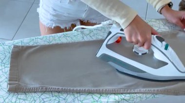 Parlak ışıklandırmayla desenli bir tahtada pantolonunu ütüleyen, modern bir ortamda ev işlerini yapan bir insan.