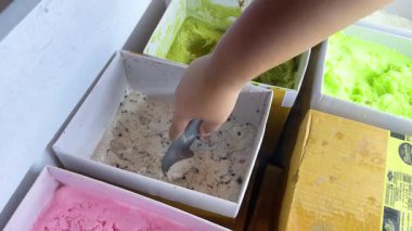Bir el, parlak aydınlatılmış bir dükkanda dondurmayı bir kaseye doldurur ve çeşitli lezzetlerin canlı renklerini ve dokularını yakalar.