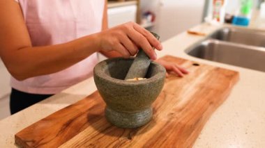 Bir insan, iyi aydınlatılmış bir mutfakta ahşap bir tahtanın üzerinde havan topu ve tokmakla malzemeleri öğütür.