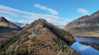 İnsansız hava aracı görüntüleri, Moke Gölü 'nün üzerinde, berrak gökyüzünün altında dağlık manzaralar ve sakin sular gösteren manzaralı bir uçuş görüntüsü yakalıyor.