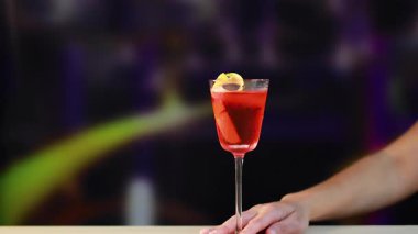 Bir el, canlı ve bulanık bir arka plana karşı zarif bir şekilde Negroni kokteyli sunar. Sahne iyi aydınlatılmış, içkiye odaklanmış.