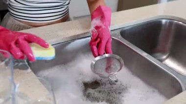 Bir insan mutfak lavabosunda pembe eldiven ve sünger kullanarak süzgeç temizler.