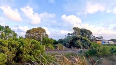 Parlak mavi gökyüzünün altında yemyeşil ve uzak evlerden oluşan sakin bir manzara Büyük Okyanus Yolu boyunca çekiliyor.