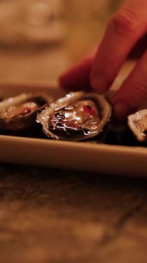 Bir el, sıcak ve loş bir ortamda bir tepsiden istiridye seçer ve samimi bir yemek atmosferi yaratır.