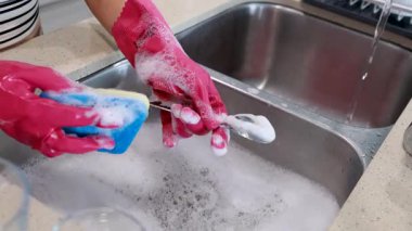 Pembe eldiven giyen biri, mutfak lavabosunda sabunlu suyla dolu bir sünger kullanarak kaşığı temizler.