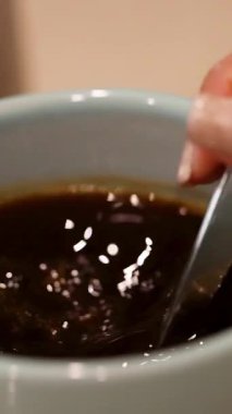 Bir el mavi bardakta kahve karıştırıyor, dalgalar ve kabarcıklar yaratıyor. Ilık aydınlatma sıcak atmosferi güçlendirir